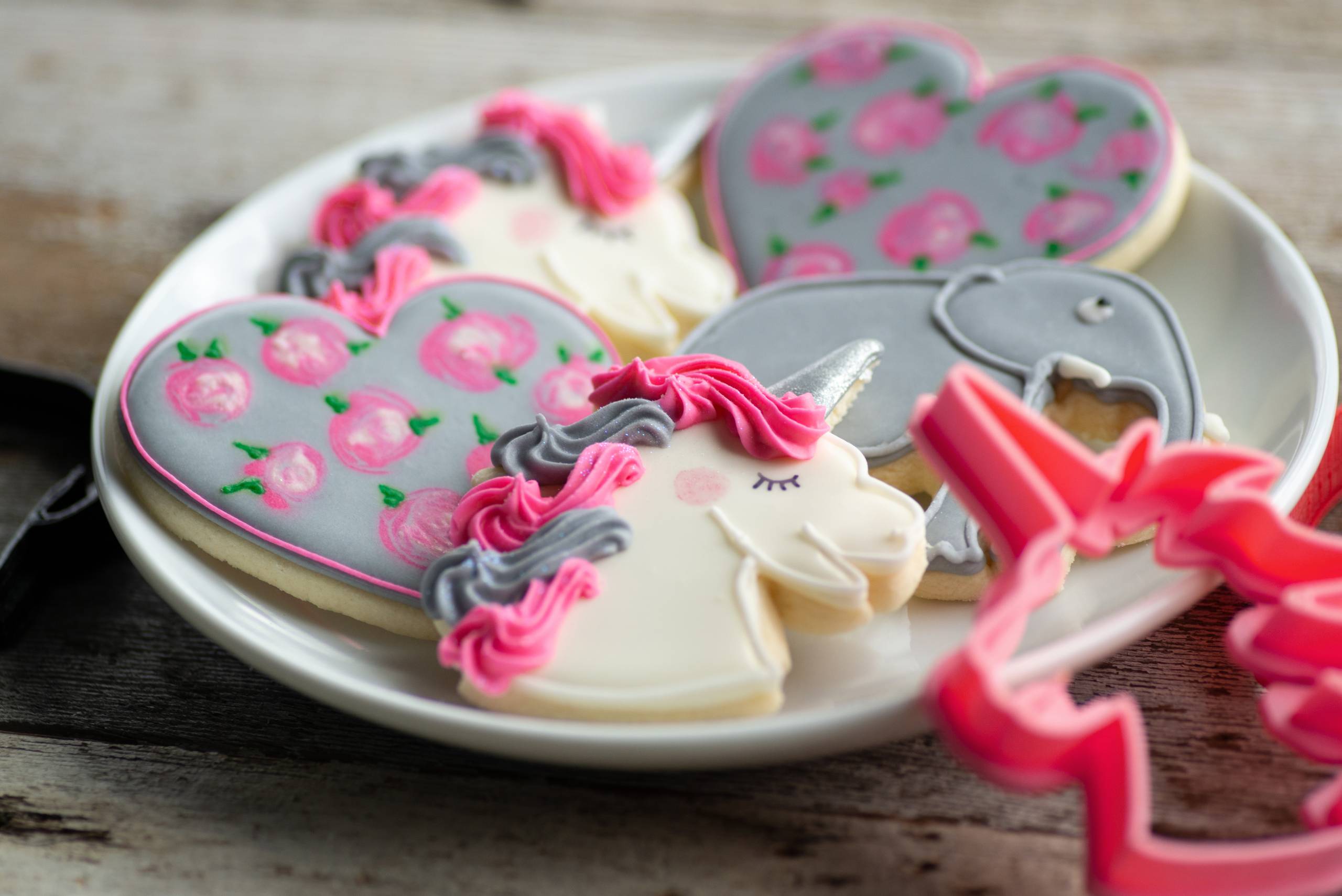 A plate of decorated sugar cookies in the shapes of unicorns and hearts with pink and gray icing. A pink unicorn-shaped cookie cutter is placed beside the plate on a wooden surface.