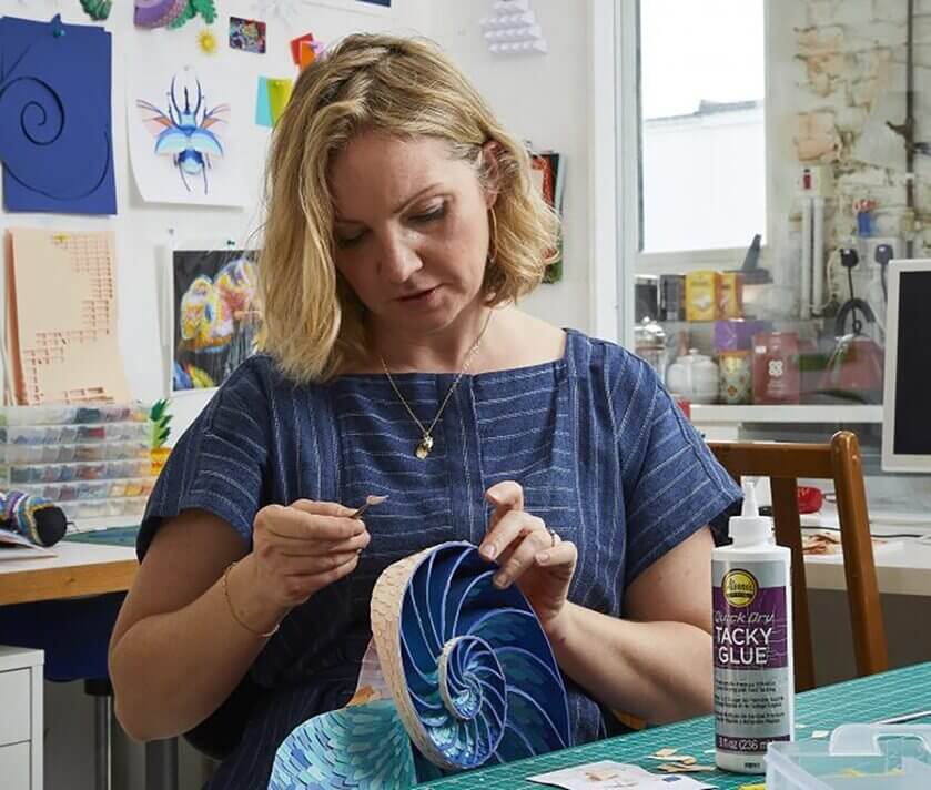 A woman in a blue dress working on an intricate paper art project in a craft room, with various art supplies and tools around her. A bottle of tacky glue is visible on the table.