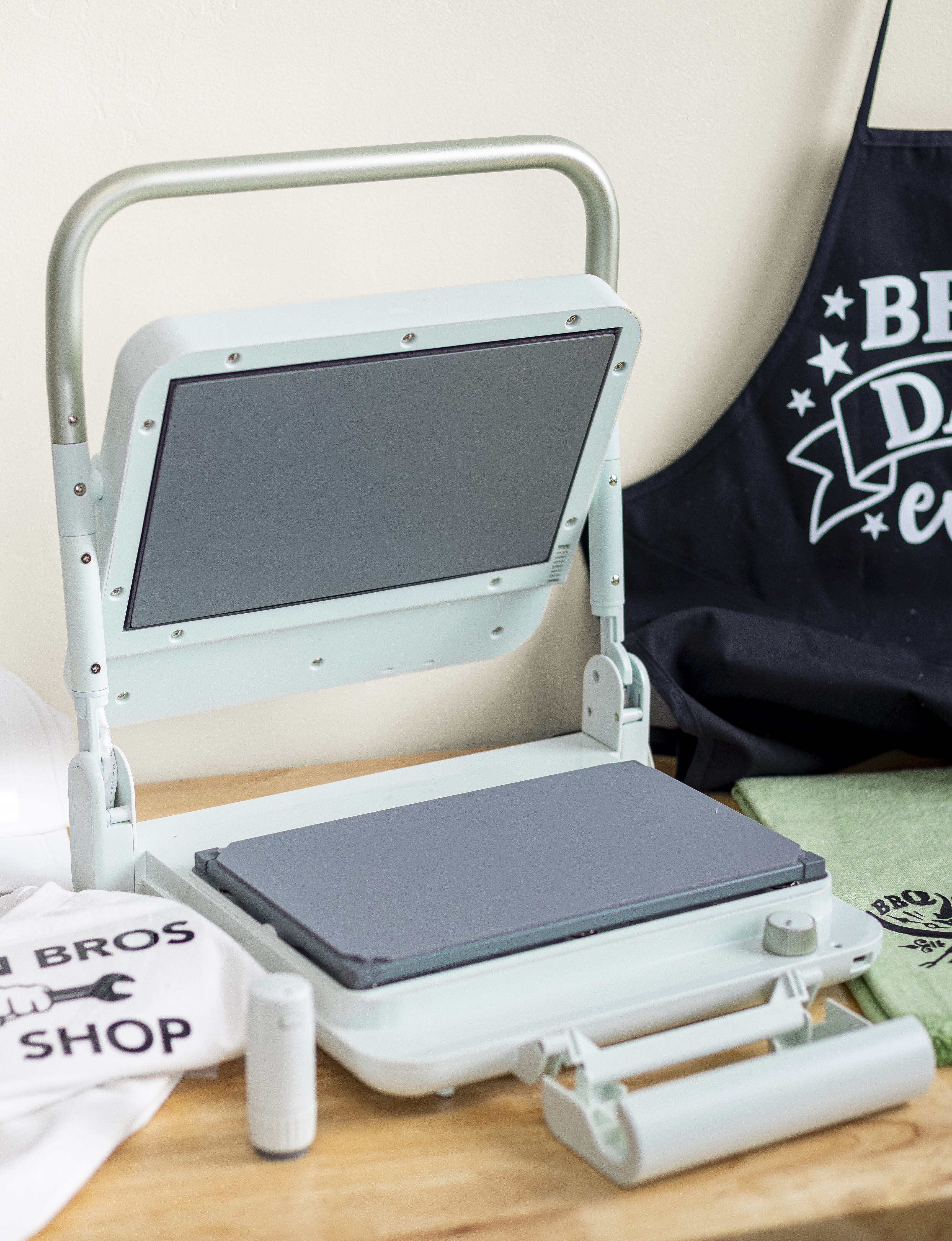 Silhouette Cloché heat press on a crafting table with apron and t-shirts, showcasing professional-quality heat transfer designs