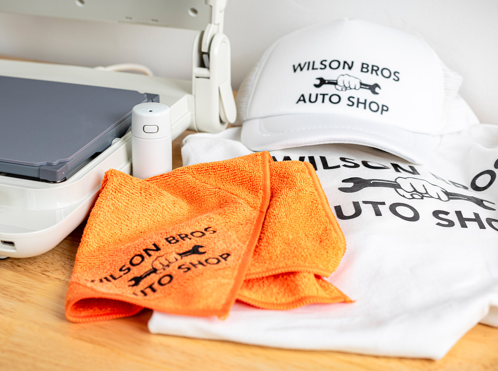 Silhouette Cloché and Petite heat presses beside custom Wilson Bros Auto Shop-branded t-shirt, cap, and microfiber cloths