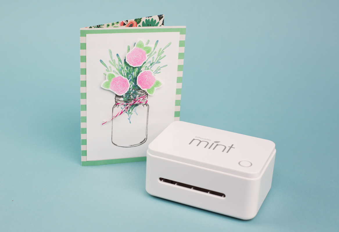 A white Silhouette Mint machine next to a handmade card featuring a bouquet of pink flowers in a jar, set against a light blue background.