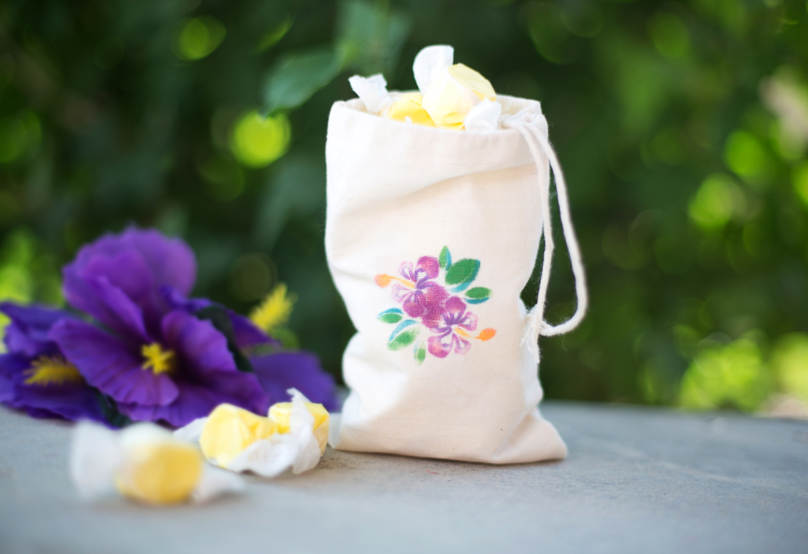 A small drawstring bag with a floral design, filled with wrapped candies, placed on a surface with purple flowers in the background. The bag has an illustration of hibiscus flowers and leaves.