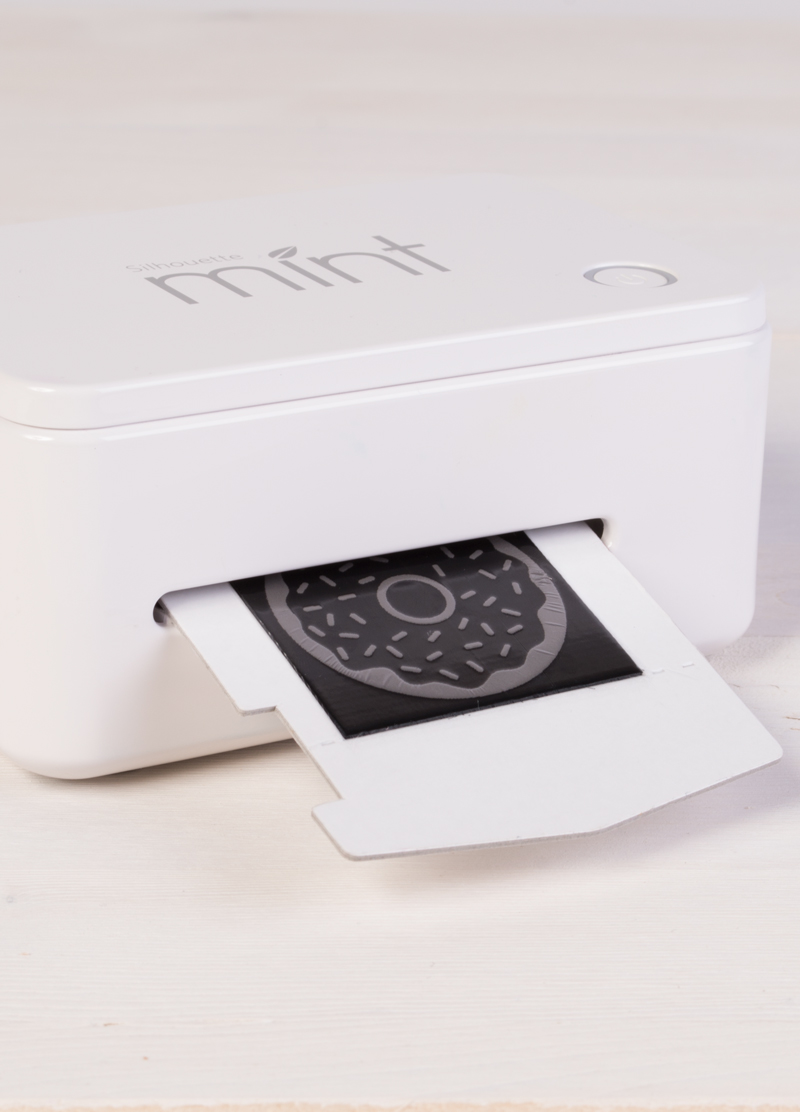 A white Silhouette Mint machine printing a stamp design of a donut with sprinkles, placed on a light wooden surface.