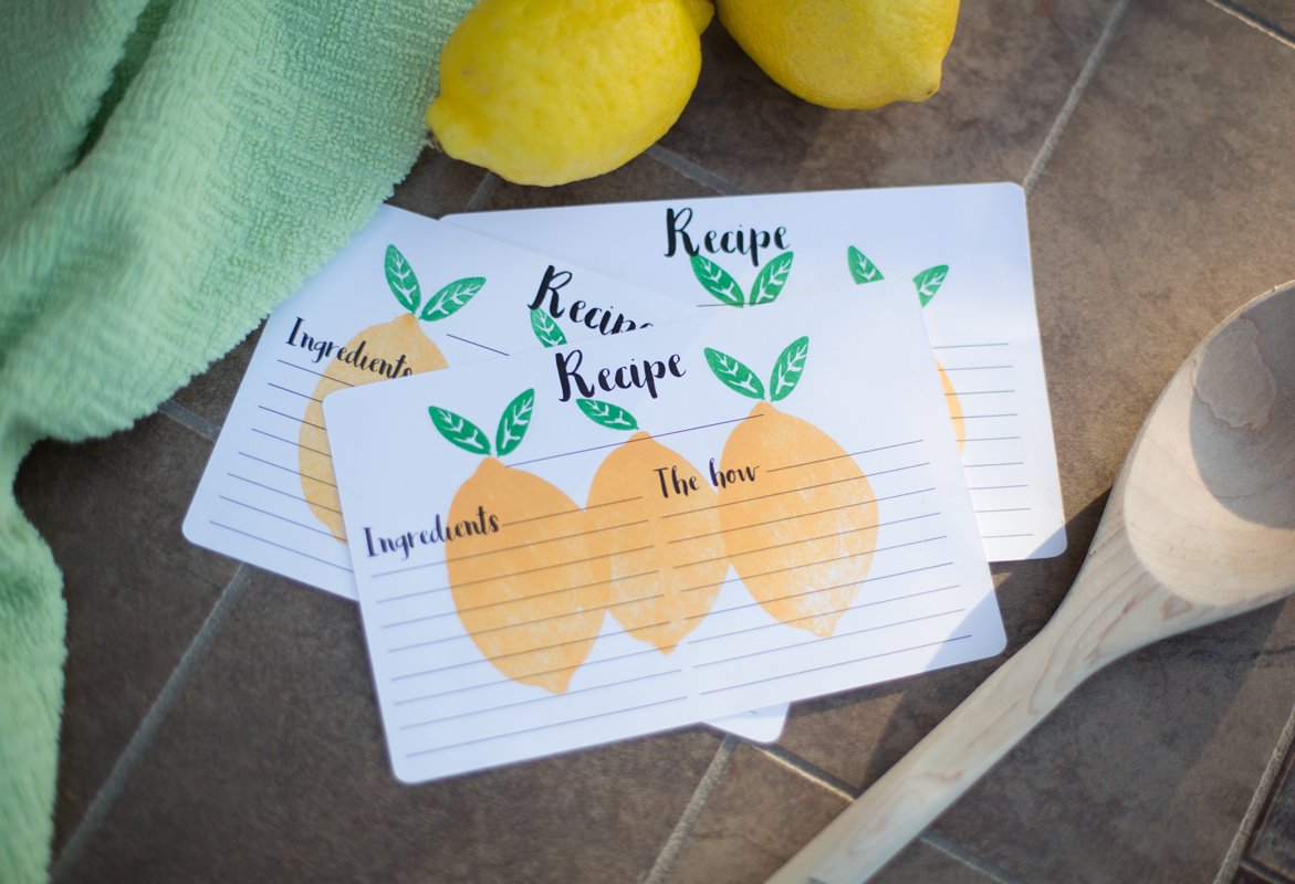 Three recipe cards with illustrations of lemons and green leaves, placed on a tiled surface next to a green towel, fresh lemons, and a wooden spoon. The cards have sections labeled 'Ingredients' and 'Recipe' or 'The how'.