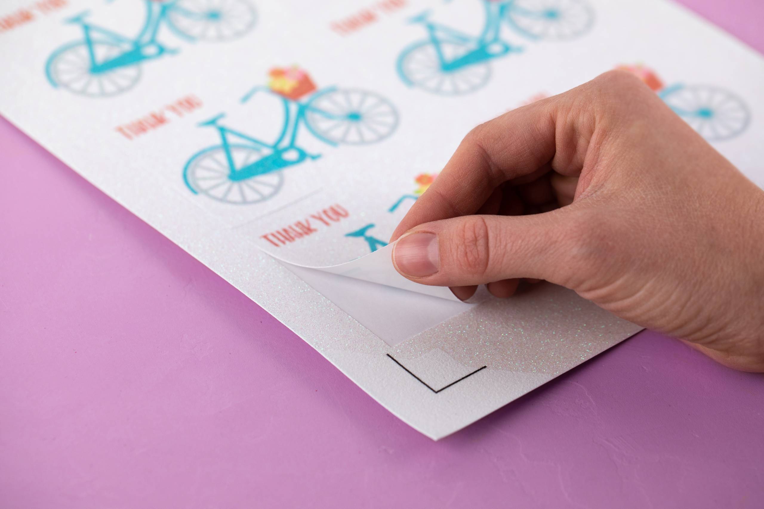Close-up of a person's hand peeling a sticker with a blue bicycle design and 'Thank You' text from a sheet of stickers on a pink background.