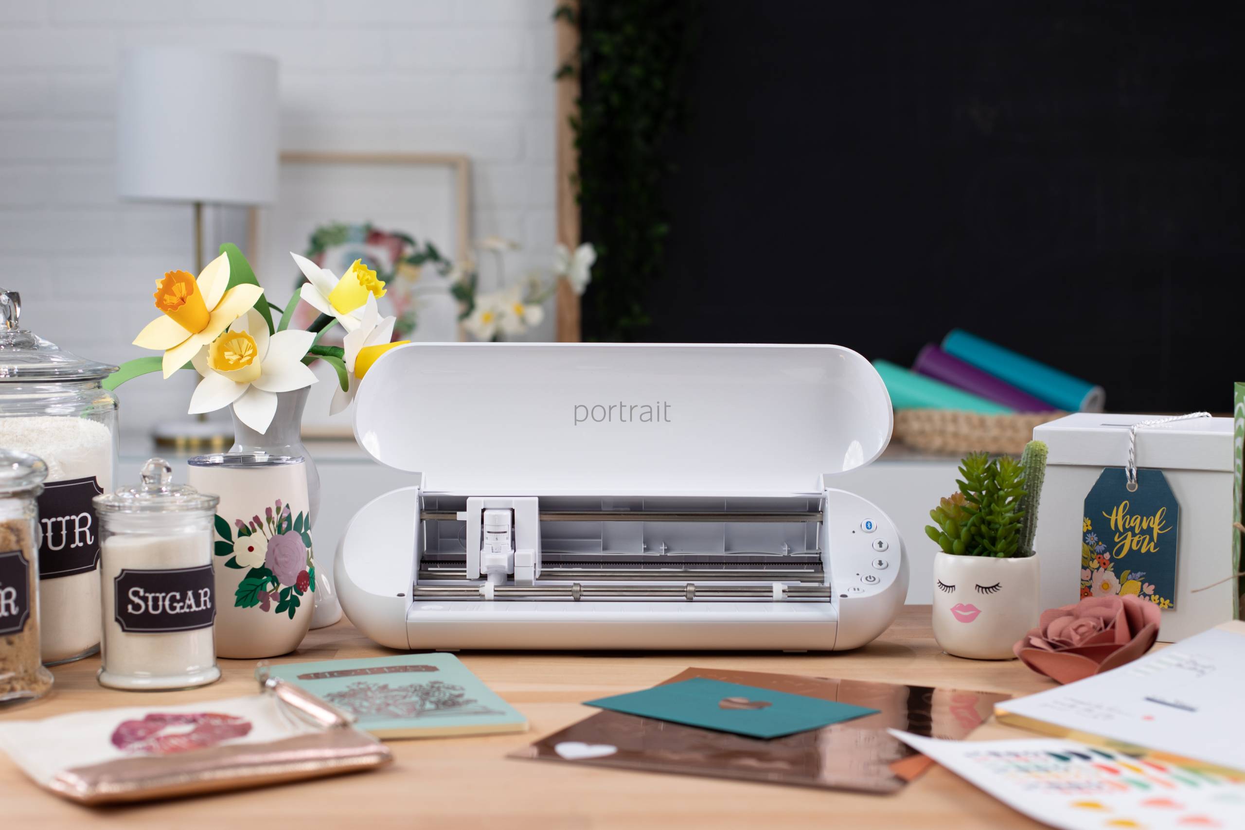 Portrait cutting machine on a table surrounded by various craft supplies, including paper flowers in a vase, labeled jars, notebooks, and a potted plant, with a thank you card and other craft projects nearby.