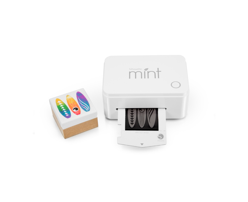 A white Silhouette Mint machine printing a stamp with surfboard designs. Next to the machine is a completed stamp with colorful surfboard illustrations mounted on a wooden block