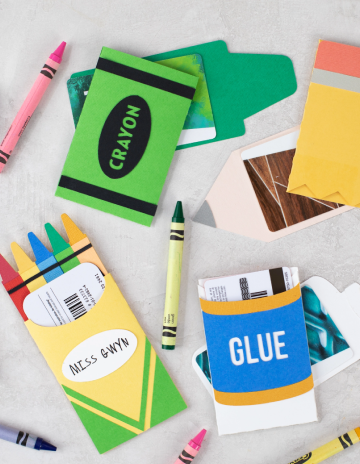 Assorted paper craft items resembling school supplies, including crayon and pencil shapes, spread out on a gray surface. Some pieces resemble crayons in various colors, while others are designed to look like crayon boxes and pencils.