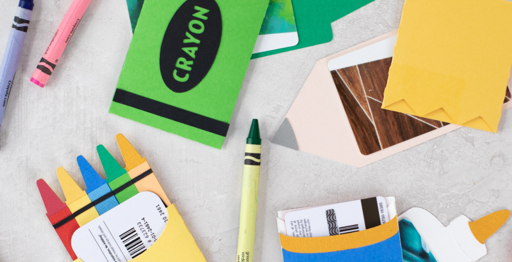 Assorted paper craft items resembling school supplies, including crayon and pencil shapes, spread out on a gray surface. Some pieces resemble crayons in various colors, while others are designed to look like crayon boxes and pencils.
