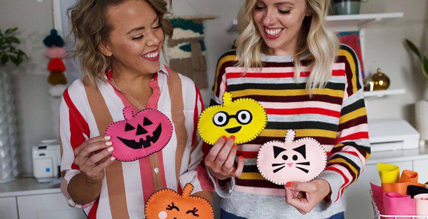 Two women smiling and holding up handmade felt pumpkins with various faces in a craft room. One woman is wearing a red and white striped shirt, and the other is wearing a multicolored striped sweater. The room is decorated with plants and craft supplies